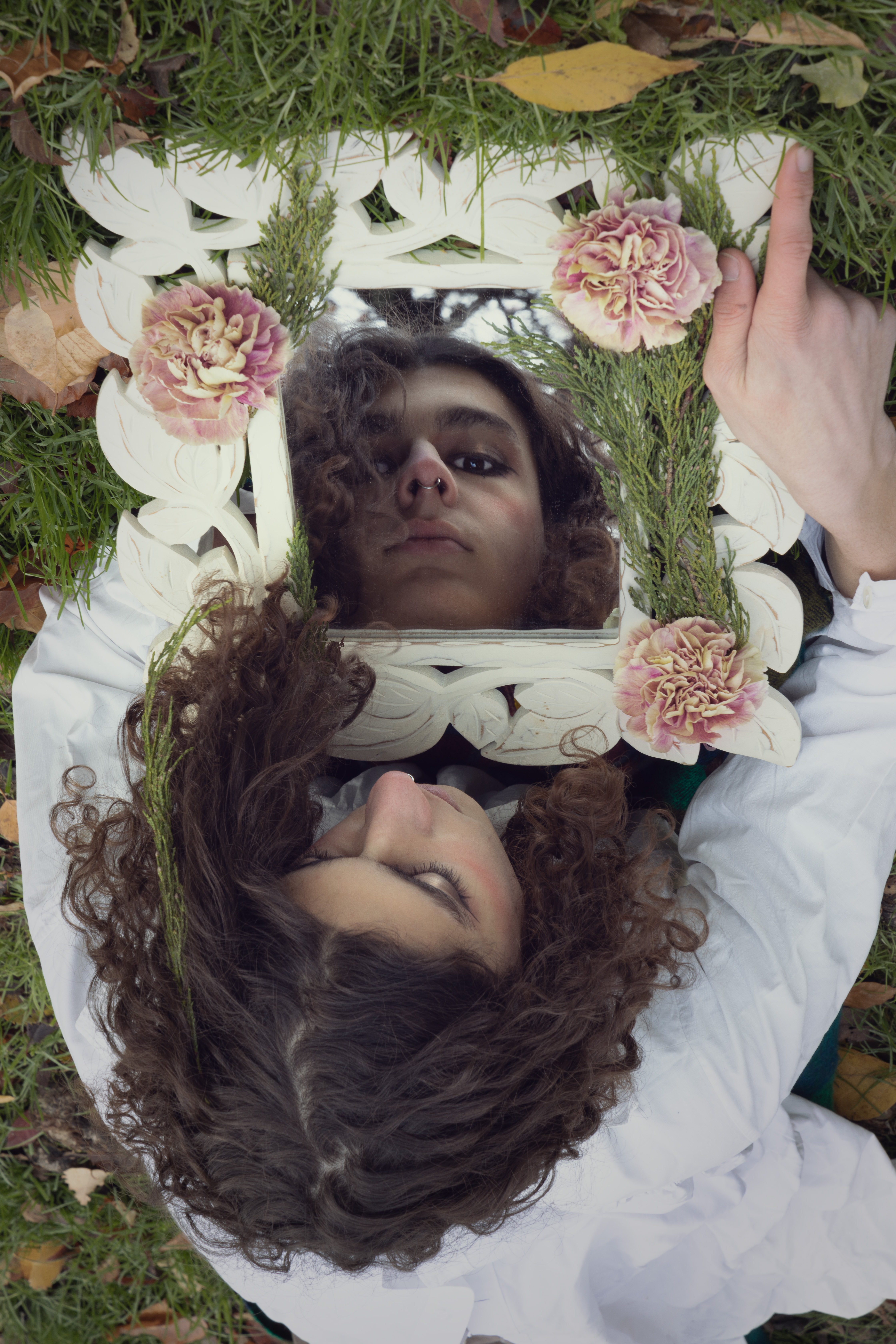Girl looking at a mirror, her reflection is looking at you, the mirror's border and her hair is decorated with mauve carnations and cypress leaves.